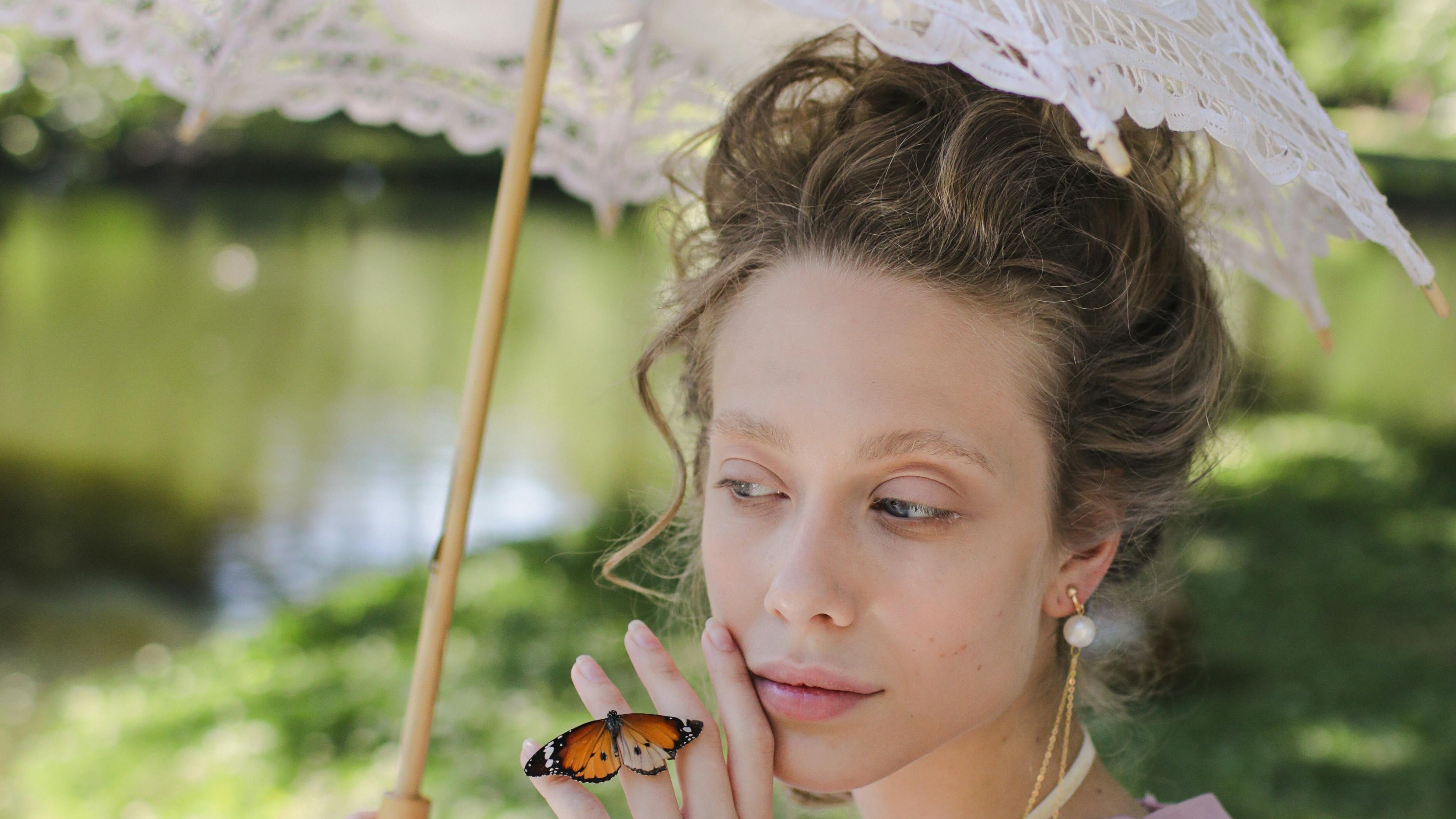 Model wearing a blush pink dress outdoors with a butterfly resting on her hand, introducing the Angélisse Beauty Crowned Collection.